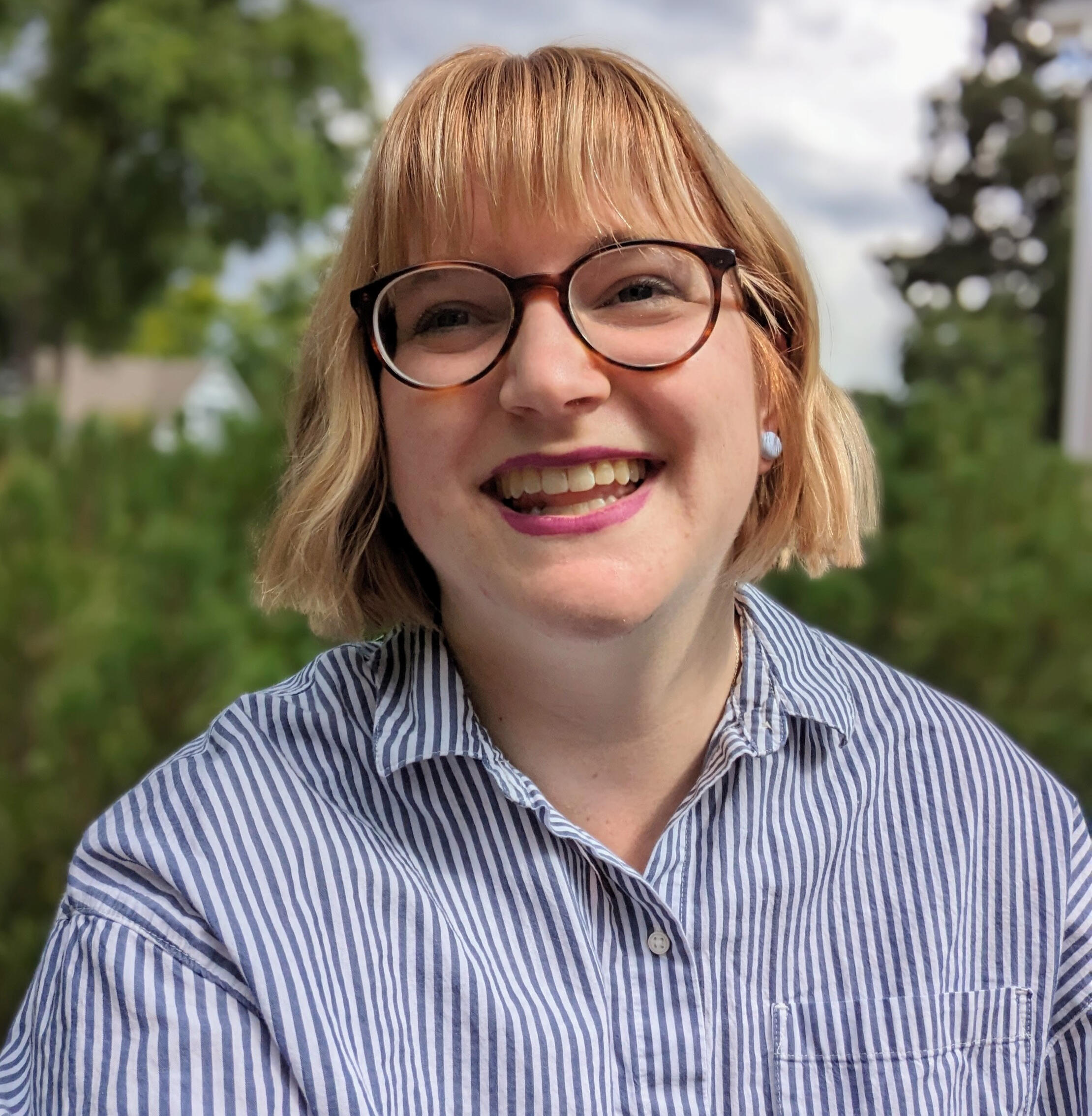 Photo of Chelsea, a white woman wearing glasses with blonde hair in a bob and bangs. She's wearing a blue-striped collared shirt and laughing as she looks to the left of the camera.
